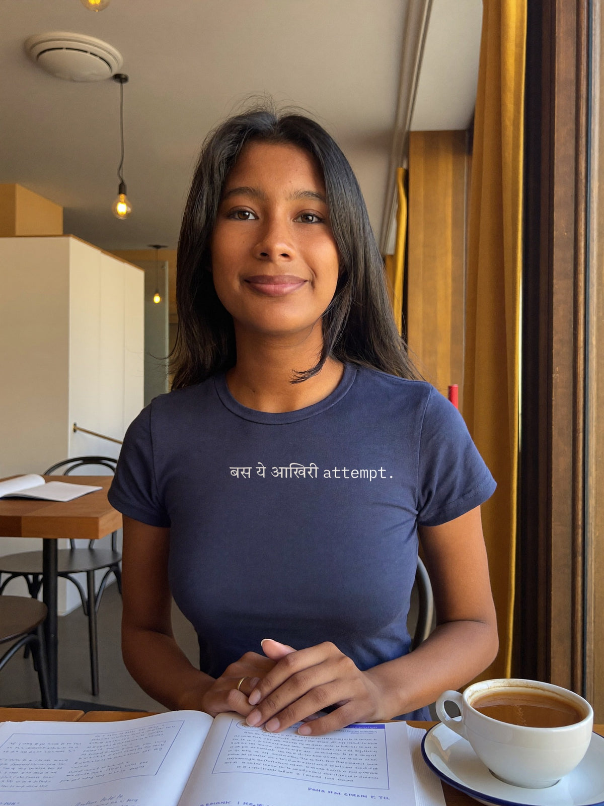 Woman wearing the “बस ये आखिरी attempt.” Hindi text t-shirt in navy by Halant, cafe