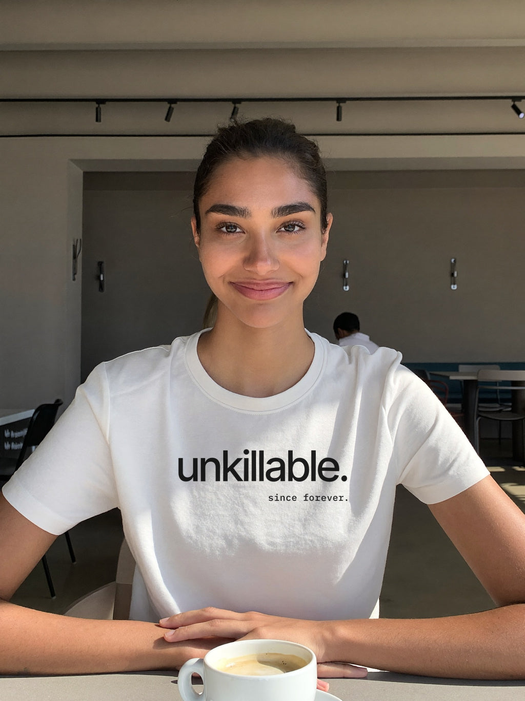 Woman wearing the “unkillable. since forever.” minimal typography t-shirt in white by Halant, cafe